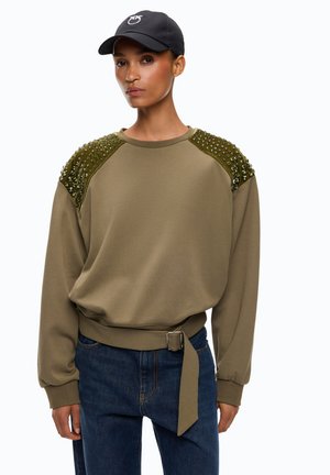Donna che indossa una felpa verde oliva con spalle borchiate, dettaglio cintura, jeans blu scuro e un berretto da baseball nero.