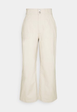 Pantalones anchos de tiro alto en color beige con bolsillos delanteros parche y cierre delantero con doble botón sobre un fondo liso.