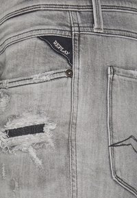 Grå slitna denimjeans med en trasig ficka och svarta stripe-detaljer. Tyget har en lätt blekt textur med synlig sömnad.