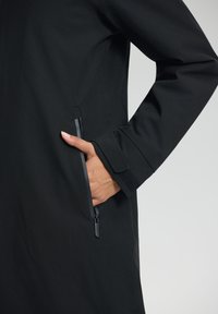 Manteau noir avec une poche latérale zippée, design sans couture et finition texturée. Les poignets incluent des accents en Velcro pour un ajustement réglable.