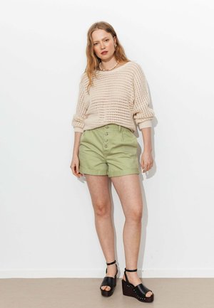 Femme debout contre un mur blanc, portant un pull en maille crème, un short taille haute vert clair, des sandales à plateforme noires et un collier en coquillage.