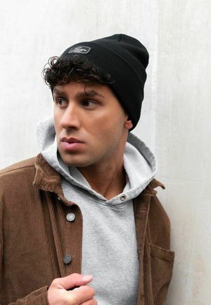 Black ribbed beanie with a logo patch, paired with a gray hoodie and brown corduroy jacket. Smooth textures and layered design details.