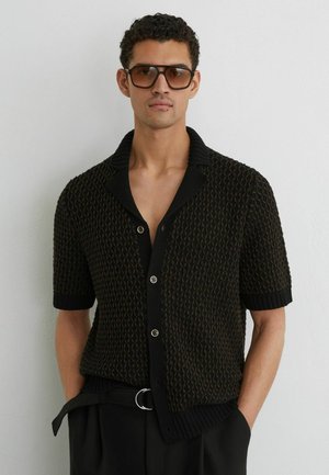 Bărbat purtând un cardigan cu mânecă scurtă, model negru, cu guler, ochelari de soare maro supradimensionați și pantaloni negri cu curea, stând în fața unui perete alb.
