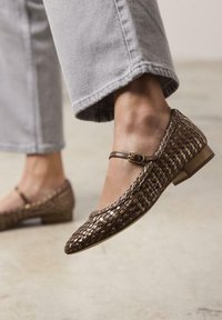 Mocasines trenzados marrones con un pequeño tacón y tira en el tobillo. El zapato presenta un diseño texturizado y una punta afilada. Se lleva con jeans grises cortos.
