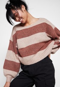 Maglione beige con righe orizzontali marroni, vestibilità over size, maniche ampie. Indossato con pantaloni neri, che mettono in mostra una texture morbida e un design rilassato.