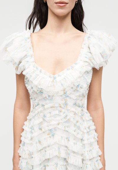 Γυναίκα που φοράει άσπρο φόρεμα με βολάν, με φουσκωτά μανίκια και διακριτικό μπλε και μπεζ floral μοτίβο, εμφανίζεται από τον λαιμό μέχρι τη μέση.