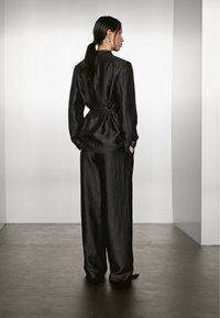 Una donna con lunghi capelli neri è di spalle, indossando un abito nero e ampio. Si trova su un pavimento liscia in uno spazio minimalista.