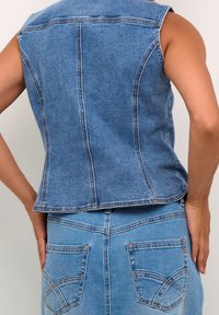 Person mit ärmellosem, tailliertem blauen Denim-Westen und hoch tailliertem blauen Denim-Rock mit Gesäßtaschen, steht gegen eine schlichte Wand.