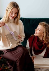 Bolsas de regalo en tonos neutros con cintas de lila; un recipiente lleva un suéter crema y una falda oscura, mientras que otro lleva una bufanda de color granate, sonriendo.