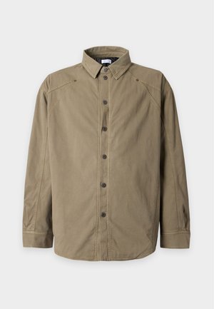 Joven con chaqueta y pantalones verdes, sudadera negra con capucha, sosteniendo una bolsa tote negra con gráfico blanco, con zapatillas blancas y grises, de pie sobre fondo blanco.