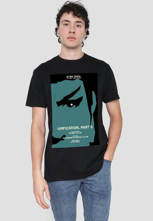 Young man wearing a black Star Trek graphic tee featuring a stylized blue face and text about "Unification, Part II" episode details.