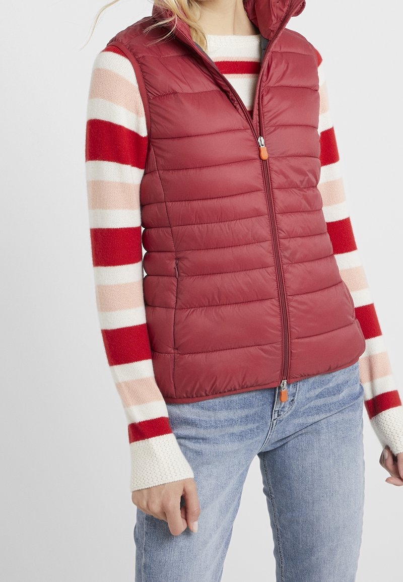 Femme portant une veste matelassée sans manches rouge par-dessus un pull rayé rouge, rose et blanc avec un jean bleu clair sur un fond blanc.