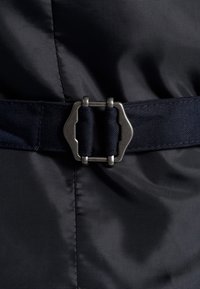 Schwarzer Stoffgürtel mit einer silbernen Metallschnalle, die eine einzigartige geometrische Form aufweist, sowie gestickten Details und strukturierten Textilakzenten.