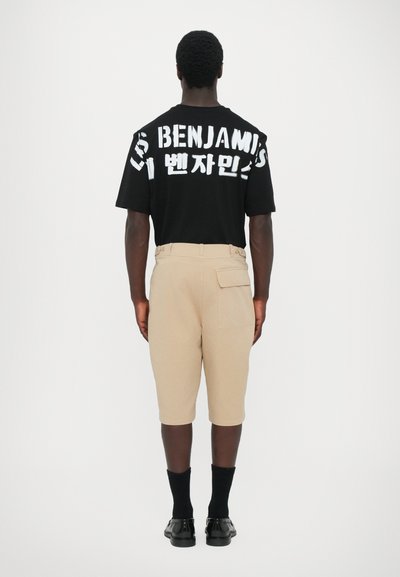 T-shirt noir à manches courtes avec du texte blanc indiquant "BENJAMINS" au dos, associé à un short beige jusqu'aux genoux et des chaussures noires.