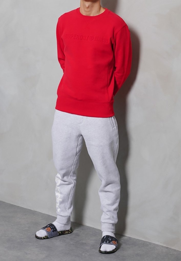 Personne portant un sweat-shirt rouge, un pantalon de survêtement gris clair, des chaussettes blanches et des sandales noires à bout ouvert, debout contre un mur gris.