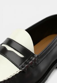 Mocassim de couro preto com um detalhe branco no peito do pé, apresentando costura decorativa e um acabamento suave. O interior tem um forro bege.