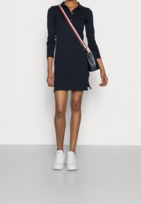 Marineblauwe polodress met lange mouwen en zijsplitten, gecombineerd met witte sportschoenen. Zwarte tas met een rood, wit en blauw hengsel.