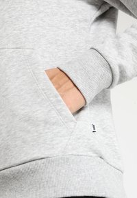 Sweatshirt gris avec poche kangourou, poignets côtelés et tissu texturé. Petite étiquette de marque visible sur la couture latérale.