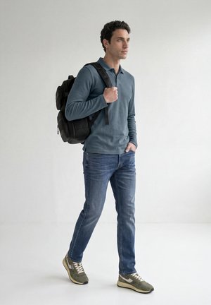 Homme marchant avec un sac à dos noir sur l'épaule, portant un polo bleu à manches longues, un jean et des baskets vertes, sur un fond clair uni.