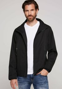 Giacca nera realizzata in materiale leggero, con zip frontale, cappuccio regolabile e due tasche laterali. Camicia bianca a scollo a V sotto.