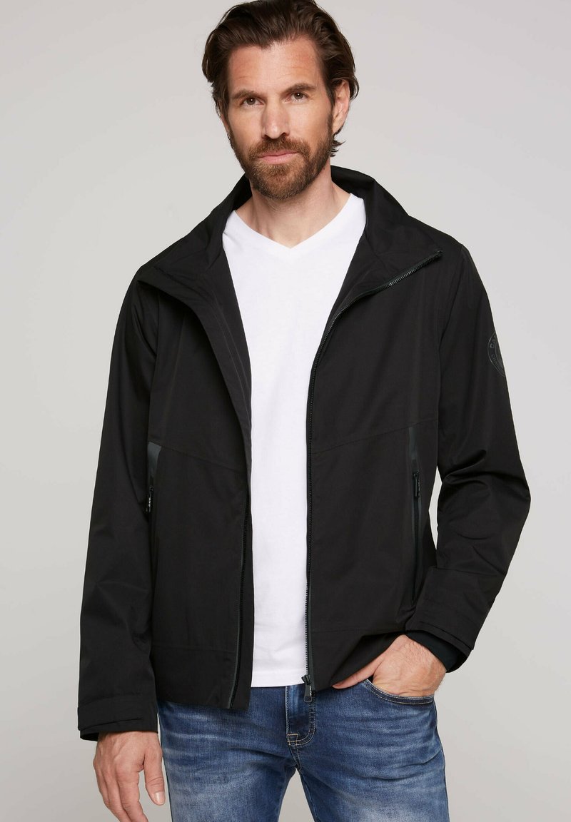 Giacca nera realizzata in materiale leggero, con zip frontale, cappuccio regolabile e due tasche laterali. Camicia bianca a scollo a V sotto.
