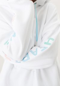 Sweat à capuche blanc avec un tissu doux, un cordon de serrage bleu clair, et un texte aux couleurs pastel sur les manches. Design simple avec une coupe confortable.
