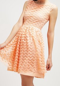 Robe couleur pêche avec un motif en zigzag, sans manches, corsage ajusté et jupe évasée, dotée d'un superposition transparente et d'une texture lisse.
