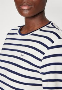 Gros plan d'une personne portant une chemise blanche et bleu marine à rayures horizontales avec "GANT" brodé sur la poitrine.