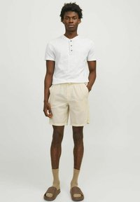 Fehér, rövid ujjú henley póló, három gombbal, bézs csíkos rövidnadrággal párosítva. A modellen világosbarna zoknik és barna slip-on szandálok vannak.