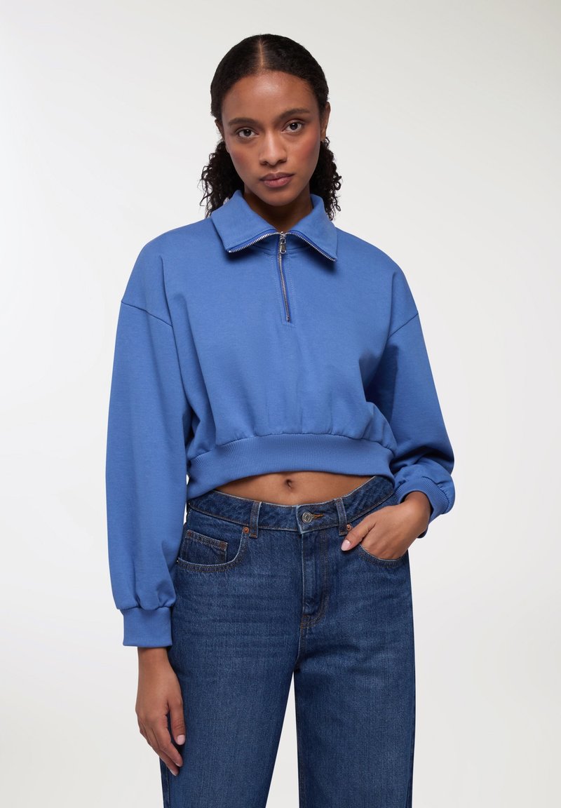 Felpa blu cropped realizzata in morbido tessuto, con colletto alto e zip frontale, abbinata a jeans alta vita di colore blu scuro.
