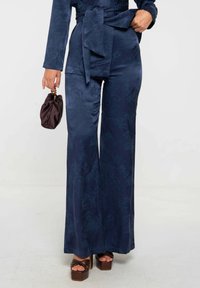 Combinaison en satin bleu marine avec des manches longues, une taille ceinturée et des jambes évasées, présentant un motif de texture subtil. Sandales à plateforme brunes.