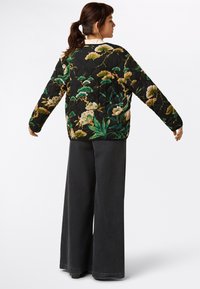 Gequilted zwarte jas met een bloemmotief in groen en roomkleur, met lange mouwen en een ontspannen pasvorm. Gecombineerd met een wijde, donkere broek.