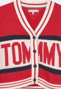 Czerwono-kremowy sweter robiony na drutach z dekoltem w serek, czarnymi guzikami i logo "TOMMY" w pogrubionej czcionce. Poziome paski w kolorze granatowym i czerwonym.