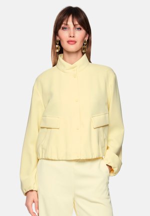 Frau mit glatten braunen Haaren, die eine blassgelbe Jacke mit Stehkragen und Druckknöpfen, Pattentaschen, passende Hose und goldene Statement-Ohrringe trägt.