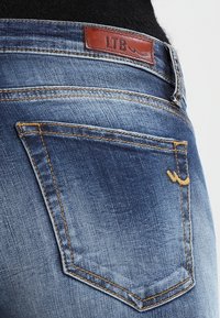 Los jeans de mezclilla azul presentan un diseño de tiro medio, con una etiqueta de cuero marrón en la cintura y costuras amarillas en el bolsillo trasero.