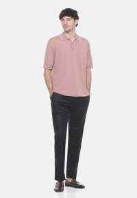 Uomo in piedi con le mani in tasca, che indossa un polo rosa polveroso a maniche corte, pantaloni neri e mocassini marrone scuro su sfondo bianco.
