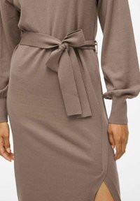 Robe en tricot taupe à manches longues et ceinture nouée. Dotée d'une fente latérale et de poignets côtelés, mettant en avant une silhouette ajustée.