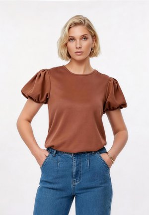 Νεαρή γυναίκα με κοντά ξανθά μαλλιά, φοράει ένα καφέ τοπ με φουσκωτά μανίκια και μπλε τζιν ψηλόμεσα, στέκεται με τα χέρια στις τσέπες.