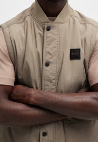 Beige bezrukávová bunda s žebrovaným límcem, cvočky a černou textilní nášivkou. Nosená přes světle růžovou košili s krátkým rukávem.