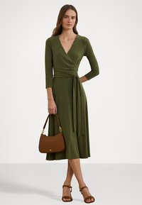 Abito avvolgente verde oliva con maniche a tre quarti e vita legata, abbinato a una borsa marrone e sandali con tacco marroni.