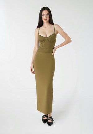 Donna con lunghi capelli scuri che indossa un abito maxi aderente verde oliva senza maniche e scarpe nere a punta con cinturini, in piedi su sfondo bianco.
