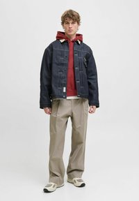 Veste en denim sombre avec un col beige, superposée sur un hoodie rouge. Un pantalon beige ample et des baskets de couleur claire complètent la tenue.