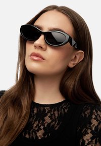Woman with long brown hair wearing black oval sunglasses and a black lace top, facing slightly upward against a white background.