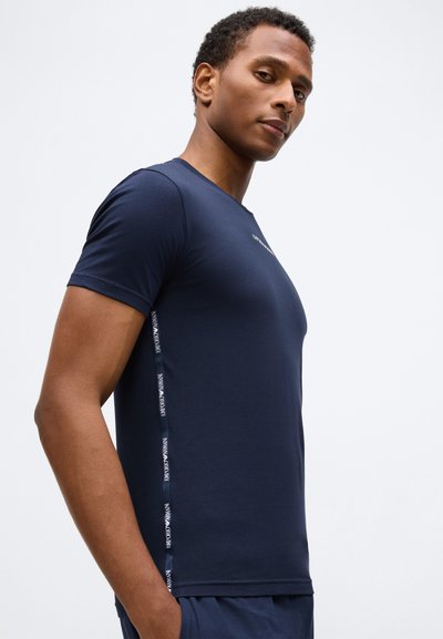 Jauns vīrietis, tērpts pieguļošā tumši zilā T-kreklā ar Emporio Armani logo lentu gar malām, pozējot uz balta fona.