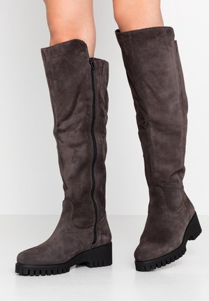 Brązowe zamszowe kozaki do kolan z czarnymi masywnymi podeszwami i bocznymi zamkami, noszone na gołych nogach na białym tle.