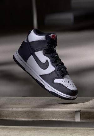 Sort og hvid Nike high-top sneakers hængende over en betongulv med snørebånd bundet og rød logo på tungen.