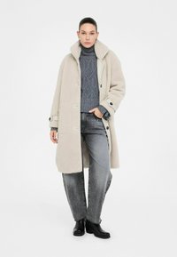 Donna che indossa un cappotto di lana beige sopra un maglione grigio a collo alto lavorato a trecce, jeans grigi slavati e stivaletti neri, in piedi su uno sfondo bianco.
