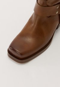 Bottine en cuir marron avec un bout carré, sangle latérale ornée de clous métalliques et texture lisse. Semelle robuste avec des détails subtils.