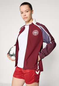 Maroon athletic jacket with striped sleeves, white zip, logo on chest; paired with white t-shirt and red shorts featuring white accents.