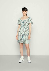 Jovem mulher com cabelo escuro veste um vestido curto floral verde menta e tênis brancos de cano alto, de pé contra um fundo claro e liso.